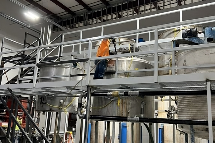 Industrial maintenance: Worker performing maintenance on large silver tanks in an industrial interior with metal scaffolding and pipes.
