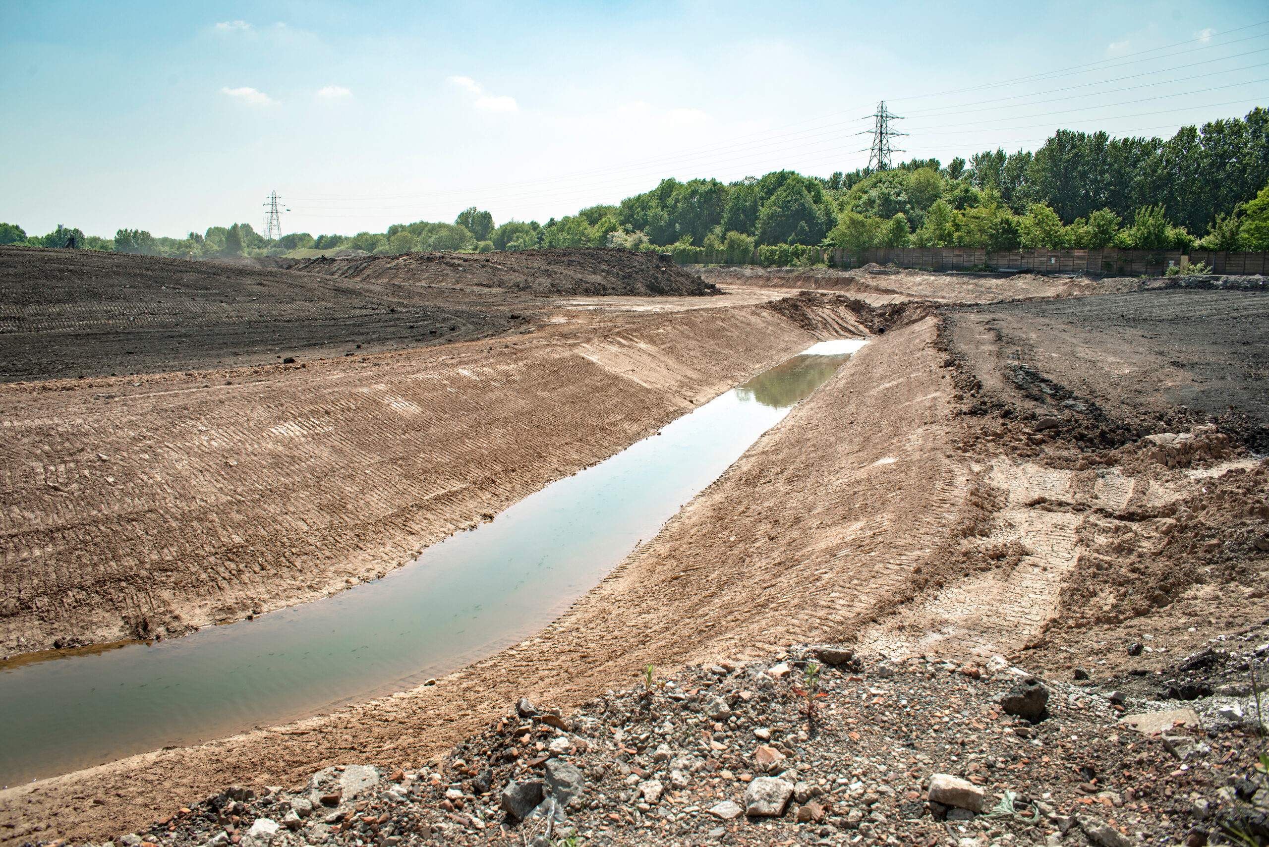 Environmental remediation: Large excavated area with a narrow channel of water, rough ground, trees and power lines in the background, showing site preparation or soil removal.