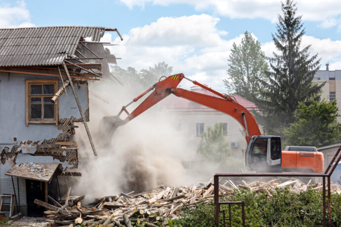 exterior demolition: in progress: orange excavator demolishing a building with a damaged roof, with dust and debris visible at the demolition site.
